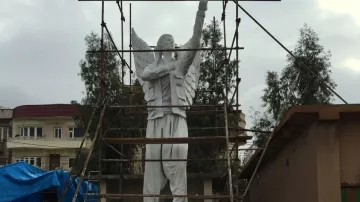 تظهر في الصورة تمثال عملاق لشخص يحمل ذراعه إلى الأعلى، يبدو أنه في مرحلة الإنشاء أو التجديد، حيث توجد scaffoldings (سقالات) تدعمه. يحيط بالتمثال بعض الأشجار والمباني. كما يوجد سطح غير مستوٍ في الأسفل وبعض المواد والأدوات المستخدمة في البناء. يناقض شكل التمثال البيئات المحيطة به، مما يبرز هيكله المميز. السماء تبدو غائمة، مما يعطي شعورًا بالتغير في الطقس.