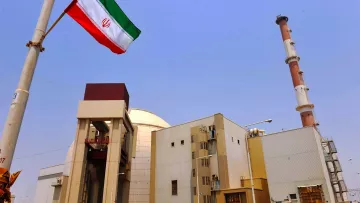 L'image montre une installation industrielle avec des structures modernes. On peut voir une grande cheminée au fond et un bâtiment principal qui semble être lié à l'énergie. Au premier plan, il y a un mât avec le drapeau iranien flottant, qui arbore des bandes rouges, blanches et vertes. Le ciel est dégagé, créant un contraste avec les bâtiments. L'ensemble évoque une infrastructure liée à la production d'énergie, probablement nucléaire.