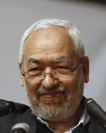 Je ne sais pas qui est cette personne, mais je peux décrire l'image de manière générale. Il s'agit d'un homme d'âge mûr avec des cheveux grisonnants et une barbe. Il porte des lunettes et a un sourire serein. L'arrière-plan semble être flou, ce qui attire l'attention sur lui. Les microphones à proximité suggèrent qu'il pourrait être en train de parler lors d'un événement ou d'une conférence.