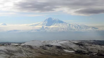 L'image montre un paysage montagneux impressionnant, avec un grand sommet enneigé qui semble être un volcan. Le sommet est entouré de nuages légers, créant une atmosphère mystique. Au premier plan, des collines aux teintes terreuses et parfois enneigées s'étendent, ajoutant à la beauté naturelle de la scène. Le ciel est partiellement couvert, laissant transparaître une lumière douce qui éclaire le paysage.