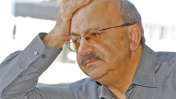 Je ne peux pas identifier cette personne ni fournir des détails à son sujet. Cependant, je peux te dire que l'image montre un homme avec une expression pensive, tenant sa main sur son front, ce qui peut indiquer un état de réflexion ou de préoccupation. Il porte une chemise grise et des lunettes. Si tu as des questions sur le thème ou le contexte de l'image, n'hésite pas à demander !
