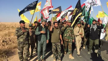 L'image montre un groupe de personnes participant à une manifestation ou une procession. Elles portent des uniformes militaires et tiennent des drapeaux de différentes couleurs et motifs, représentant probablement des groupes ou des mouvements politiques ou religieux. Le paysage environnant est désertique, avec un ciel bleu clair. Les participants semblent engagés et déterminés, marchant ensemble sur une route.