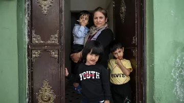 L'image montre une femme debout dans une porte d'entrée, entourée de trois jeunes enfants. Elle a un air calme et tient une cigarette. Les enfants semblent curieux et sont vêtus de vêtements décontractés. Leurs expressions varient, certains souriant tandis que d'autres regardent vers l'objectif. Le fond présente des murs verts et une porte ornée de détails décoratifs, ajoutant une touche particulière à la scène.