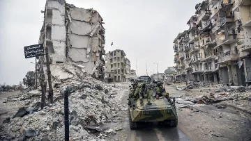 L'image montre une scène de destruction urbaine, probablement à la suite d'un conflit. On peut voir des bâtiments en ruine, avec des façades effondrées et des débris éparpillés sur le sol. Au premier plan, il y a un véhicule militaire, sur lequel des soldats sont présents. L'atmosphère semble sombre et chargée, soulignant les ravages causés par la guerre.