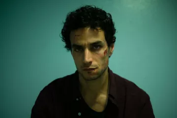L'image montre un homme assis devant un mur de couleur bleu clair. Il a des cheveux noirs et bouclés, et son expression faciale semble intense ou préoccupée. Son visage présente des éraflures ou des blessures visibles, ce qui suggère qu'il a traversé une situation difficile. Il porte une chemise foncée et a l'air fatigué ou en détresse. L'éclairage est dramatique, mettant en avant ses traits et les blessures sur son visage.