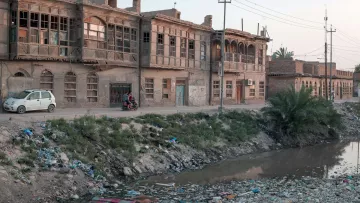 L'image montre une rue bordée de vieilles maisons en briques, caractérisées par des fenêtres en bois. À gauche, on aperçoit une voiture garée. Sur le côté droit, un groupe de personnes semble discuter. Au premier plan, un bras de rivière est visible, rempli de déchets et de débris, entouré de végétation. L'éclairage doux de la fin de journée crée une atmosphère nostalgique dans ce paysage urbain en dégradation.