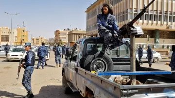 تظهر في هذه الصورة مجموعة من عناصر الأمن أو الشرطة، يرتدون زيًا رسميًا باللون الأزرق، بينما يقومون بالتحرك في منطقة حضرية. أحدهم يجلس على الجزء الخلفي من شاحنة صغيرة، حيث يوجد مدفع ثقيل مثبت عليها. هناك سيارات أخرى في الخلفية ومباني كبيرة على الجانبين. تعكس الصورة أجواء من النشاط الأمني أو الاستعداد للتعامل مع موقف معين.