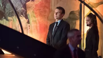 L'image montre un homme en costume, debout sur une scène, avec une femme à ses côtés qui porte un microphone et un ouïe. L'arrière-plan présente des couleurs et des formes abstraites, suggérant un environnement artistique ou un événement important. L'homme semble attentif, tandis que la femme semble concentrée sur la situation. Il y a également un autre homme en arrière-plan qui marche, suggérant une ambiance dynamique.