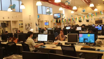 L'image montre un environnement de bureau moderne et animé. Plusieurs employés sont assis devant des ordinateurs, travaillant sur leurs tâches. Les bureaux sont équipés de plusieurs écrans d'ordinateur. La pièce est décorée avec des ballons et des banderoles, suggérant une atmosphère festive ou un événement spécial. La lumière naturelle entre par de grandes fenêtres, ajoutant une ambiance agréable à l'espace de travail.