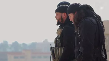 تظهر الصورة مجموعة من الرجال يرتدون ملابس داكنة. يبدو أنهم في وضعية استعداد، حيث يمكن رؤية بعضهم من الجانب. يرتدي البعض منهم شارات أو معدات تكتيكية، مما يدل على أنهم قد يكونون في سياق عمل يتعلق بالأمن أو العمليات العسكرية. خلفهم يظهر مشهد غير واضح، لكن يمكن رؤية بعض المباني في الخلفية. الأجواء عامة تبدو جادة.