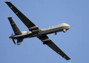 L'image montre un drone de type militaire, en vol. Il a une conception aérodynamique avec des ailes étendues et une queue distinctive. La surface du drone est peinte en gris, ce qui le rend moins visible en altitude. On peut également observer des équipements sous ses ailes, probablement destinés à la surveillance ou à des missions de reconnaissance. Le ciel en arrière-plan est dégagé, mettant en valeur le drone en mouvement.