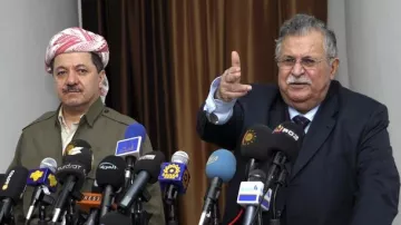 L'image montre deux hommes en conférence de presse, entourés de plusieurs micros et caméras provenant de divers médias. L'homme à gauche porte une tenue traditionnelle kurde avec un foulard, tandis que l'homme à droite, habillé en costume, semble s'adresser à l'audience en pointant du doigt. L'ambiance est formelle, indiquant un moment important de communication ou d'annonce.