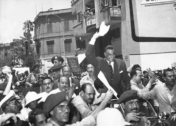 L'image montre une scène de foule animée, probablement lors d'un événement historique. De nombreuses personnes sont rassemblées autour d'une voiture, où un homme, visiblement en position d'autorité, lève les bras en signe de salutation ou d'approbation. Les spectateurs semblent enthousiastes, agitant des banderoles ou des papiers. On peut voir des policiers et des militaires en uniforme, ajoutant à l'ambiance de la scène. L'architecture des bâtiments en arrière-plan suggère une ville dynamique, probablement dans un contexte politique ou social significatif.