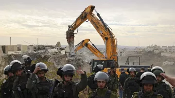 The image depicts a scene of destruction where a construction or demolition vehicle is actively tearing down a building, surrounded by a group of armed soldiers. The soldiers are wearing helmets and tactical gear, indicating a heightened security context. The background features a landscape with additional buildings, suggesting a possibly urban or conflict-affected area. The atmosphere appears tense, reflecting the serious nature of the situation being portrayed.