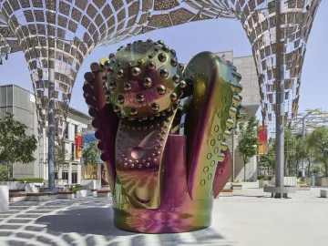 L'image présente une sculpture moderne au design futuriste, composée de formes organiques et géométriques. Les surfaces de la sculpture sont brillantes et colorées, avec des reflets irisés qui passent du vert au violet. Elle semble être placée sous une structure architecturale légère, qui offre de l'ombre et favorise une ambiance artistique. L'environnement autour de la sculpture est minimaliste, avec des arbres et quelques assises, ce qui met en valeur l'œuvre et crée une atmosphère apaisante.