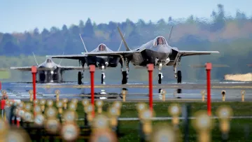 The image features three F-35 fighter jets on an airstrip, showcasing their sleek and modern designs. The jets are positioned closely together, and the background features a blurred green landscape, indicating a rural setting. The foreground includes several lights lining the runway, which adds a sense of depth to the scene. The overall atmosphere appears dynamic and focused on military aviation.