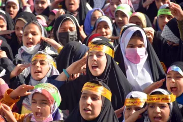 The image shows a large group of girls and young women gathered together, likely participating in an event or demonstration. They are wearing colorful headscarves and black clothing, along with yellow headbands that feature Arabic text. The girls appear to be actively engaged, singing or chanting, and expressing enthusiasm. Their expressions convey a sense of unity and purpose as they participate in this collective activity.