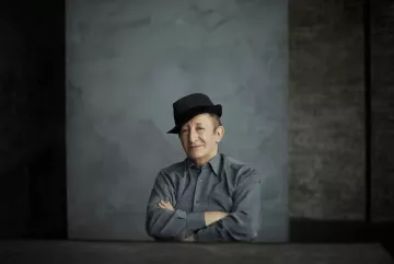 L'image montre un homme assis avec un chapeau noir. Il a un léger sourire et les bras croisés sur une table. Il porte une chemise grise. L'arrière-plan est simple, avec une texture de mur gris qui crée une atmosphère sobre et contemplative. La lumière est douce, accentuant les traits de son visage.