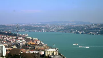L'image montre un panorama d'une ville au bord d'une grande étendue d'eau. On aperçoit un pont moderne qui traverse la rivière, reliant les deux rives. À gauche, des bâtiments s'échelonnent sur la colline, tandis qu'à droite, on peut voir des constructions historiques. Le ciel est clair, et l'ensemble du paysage évoque une atmosphère tranquille et pittoresque. On distingue également des bateaux naviguant sur la rivière.