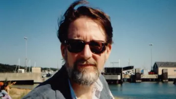 Je ne sais pas qui est cette personne, mais je peux décrire l'image. On y voit un homme avec des lunettes de soleil et une barbe, assis près de l'eau. Il a les cheveux bruns et un look décontracté, portant une veste. En arrière-plan, on aperçoit des bateaux et un quai, ce qui donne une ambiance maritime. Le ciel est clair et ensoleillé.