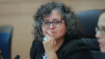 L'image montre une femme avec des cheveux bouclés et un regard réfléchi. Elle porte des lunettes et une veste. Une main est placée sur son menton, ce qui suggère qu'elle est en train de penser ou de considérer une question. L'environnement semble être une salle de réunion ou une assemblée, avec des chaises et une table en arrière-plan.
