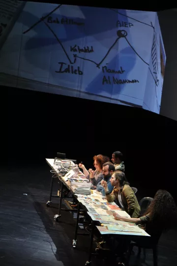 L'image montre un groupe de personnes assises autour d'une grande table sur scène, avec des ordinateurs portables et des documents éparpillés. En arrière-plan, un écran projette un schéma ou une carte, probablement liée à un contexte géographique ou politique, mentionnant des lieux comme Idleb et d'autres noms. Les participants semblent engagés dans une discussion ou une activité collaborative, ce qui suggère un travail de groupe intense. L'ambiance est dynamique, mettant en avant l'importance de l'interaction et de l'échange d'idées.