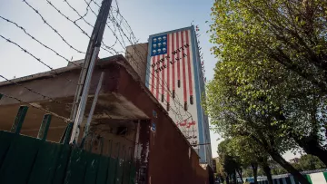 L'image montre un mur d'un bâtiment avec une grande peinture représentant un drapeau américain à rayures. Le mur est bordé par une clôture en barbelés, et on peut voir des arbres sur le côté. Le paysage est urbain, avec un chemin pavé qui suggère une rue à proximité. La composition évoque un contraste entre l'élément décoratif du drapeau et la présence de la clôture de sécurité.