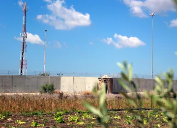 L'image montre un paysage avec un mur en béton qui semble délimiter une zone. Au fond, on peut voir des pylônes de communication et des lampadaires, suggérant une certaine infrastructure. Au premier plan, il y a un champ de cultures avec des lignes de légumes, probablement dans un état de sécheresse ou de négligence, tandis qu'un feuillage flou dans la partie inférieure de l'image ajoute une touche naturelle. Le ciel est bleu avec quelques nuages.