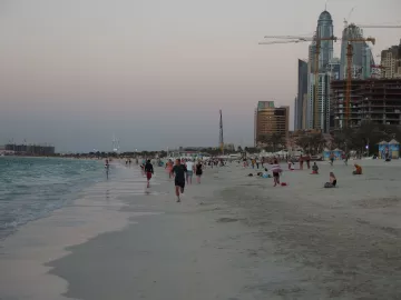 L'image montre une plage avec des personnes marchant le long du rivage. On aperçoit des baigneurs et des groupes assis sur le sable. En arrière-plan, on distingue des bâtiments modernes et des gratte-ciel, symboles d'une grande ville, ainsi que des grues témoignant de travaux de construction. Le ciel présente des teintes douces, probablement au moment du crépuscule, créant une atmosphère paisible et attractive. Les vagues de la mer semblent calmes, ajoutant à la sérénité du paysage.