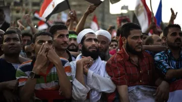 L'image montre un groupe de personnes rassemblées lors d'une manifestation. On peut voir des hommes de différentes origines, certains portant des vêtements traditionnels, d'autres des t-shirts colorés. Ils semblent tous attentifs et engagés, certains levant les mains ou tenant des drapeaux. L'ambiance semble être celle d'une mobilisation ou d'une expression de soutien pour une cause. Le fond montre un environnement urbain, avec d'autres manifestants et des drapeaux visibles.