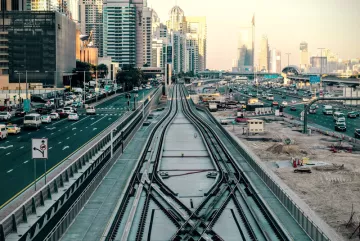 L'image montre une vue urbaine d'une ville moderne. On peut voir des routes animées avec des voitures circulant, ainsi qu'un important réseau de rails de train. À droite, il y a des bâtiments imposants et modernes qui reflètent l'architecture contemporaine. L'atmosphère est lumineuse, suggérant un moment de la journée proche du coucher du soleil avec des lumières douces. Les rails semblent bifurquer, suggérant une intersection, tandis que des travaux de construction ou d'aménagement sont visibles au premier plan.