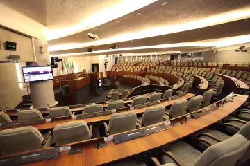 L'image montre une salle de réunion ou un hémicycle, probablement utilisée pour des sessions parlementaires ou des assemblées. On peut voir des rangées de sièges confortables disposés en arc de cercle, avec des tables en bois devant chaque siège. Des écrans et des équipements audiovisuels sont visibles, indiquant que la salle est équipée pour des présentations ou des débats. Les murs sont décorés de manière sobre et moderne, créant une ambiance institutionnelle.