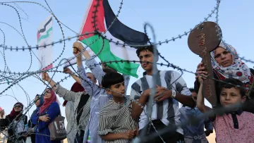L'image montre un groupe de personnes, y compris des enfants, qui tiennent des drapeaux palestiniens et des pancartes. Ils se tiennent derrière une barrière semblable à du barbelé. Les expressions des personnes peuvent communiquer des émotions telles que la détermination ou l'espoir. L'environnement semble être en plein air, sans autre détail contextuel visible.