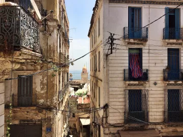 L'image montre une vue d'une rue étroite et animée d'une ville méditerranéenne. De chaque côté, on voit des bâtiments anciens avec des façades usées et des balcons en fer forgé. Des fils électriques serpentent entre les immeubles. Au loin, on peut apercevoir un dôme qui pourrait appartenir à une église ou une mosquée, et un bout de mer est visible, ajoutant une touche de bleu au paysage. Les fenêtres des bâtiments sont ornées de volets ou de rideaux, et l'ensemble dégage une atmosphère nostalgique et pittoresque.