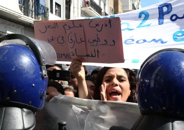 L'image montre une scène de manifestation où des personnes se rassemblent. À l'avant-plan, une femme semble exprimer des émotions vives, probablement en train de parler ou de crier, tandis qu'elle tient une pancarte. Plusieurs agents de la police, en uniforme avec des casques, se tiennent en ligne, formant un barrage. En arrière-plan, on peut apercevoir d'autres manifestants et une bannière portée par la foule. L'atmosphère semble tendue et engagée, avec une forte volonté de faire entendre des revendications.