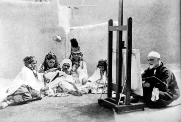 L'image montre un groupe de personnes assises autour d'un homme qui peint sur un chevalet. Les membres du groupe portent des vêtements traditionnels, certains avec des bijoux et des motifs distinctifs. L'homme qui peint semble concentré sur son travail, tandis que les autres l'observent attentivement. L'environnement paraît simple, avec des murs en argile en arrière-plan, ce qui donne un aperçu d'une ambiance culturelle et artistique.