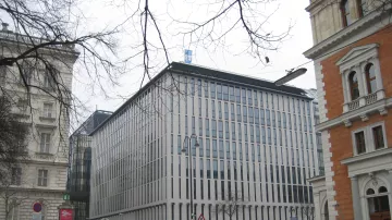 این تصویر نمای یک ساختمان مدرن را نشان می‌دهد که دارای طراحی مینیمالیستی است. نمای ساختمان با پنجره‌های عمودی و خطوط باریک تقسیم شده، ظاهری مرتب و شیک دارد. اطراف ساختمان ممکن است شامل خودروها و درختان بدون برگ در فصل زمستان باشد. در سمت چپ و راست تصویر، ساختمان‌های دیگری با طراحی‌های کلاسیک‌تر وجود دارند که تضادی جالب با ساختمان مدرن ایجاد می‌کنند. فضا به نظر می‌رسد که در یک منطقه شهری و ممکن است در فصل سرد سال باشد.