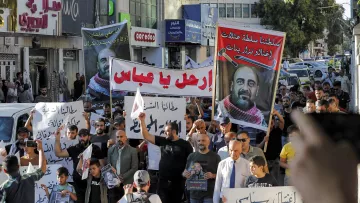 الصورة تُظهر مجموعة من المتظاهرين في شارع يجمعهم الاحتجاج، حيث يحملون لافتات كبيرة تحمل شعارات ومطالب تعبر عن قضايا معينة. يظهر في الصورة عدد من الرجال والنساء والأطفال، وبعضهم يرتدي القمصان بتصاميم مختلفة. يبدو الحشد نشطاً ومتفاعلاً، واللافتات مكتوبة باللغة العربية، مما يدل على أن التظاهر يتناول قضايا محلية. الجو يبدو حماسياً مع وجود بعض السيارات في الخلفية.