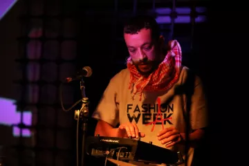 L'immagine mostra un musicista che sta esibendosi su un palco. Indossa una maglietta con la scritta "FASHION WEEK" e una sciarpa a quadri. È concentrato mentre suona un dispositivo elettronico, probabilmente un sintetizzatore o una drum machine. Il palco è illuminato da luci colorate che creano un'atmosfera vivace e coinvolgente. Dietro di lui, si intravedono proiezioni visive che aggiungono dinamismo alla performance.