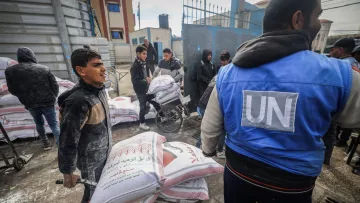 L'image montre une scène d'assistance humanitaire. Des enfants et des adultes sont occupés à transporter des sacs de nourriture, probablement du grain ou des provisions, à l'aide de brouettes. On peut voir des sacs empilés à l'arrière-plan, ainsi que des personnes portant des vêtements de protection ou des blousons. Un homme porte un gilet avec l'insigne des Nations Unies (UN), indiquant qu'il fait partie d'une mission humanitaire. L'atmosphère semble dynamique, avec un engagement collectif pour aider ceux qui en ont besoin.