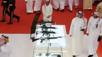 L'image montre un rassemblement où plusieurs personnes, probablement en tenue traditionnelle, se déplacent dans un espace décoré. Au centre, il y a une vitrine en verre contenant diverses armes, mettant en avant des fusils et d'autres équipements. L'ambiance semble formelle et le sol est recouvert d'un tapis rouge. Des membres de la sécurité ou des uniformes militaires peuvent être aperçus en arrière-plan, soulignant l'importance de l'événement.