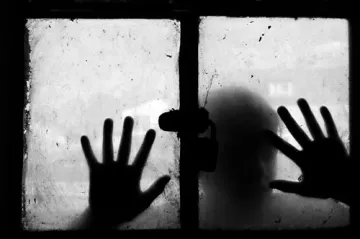 L'image montre une fenêtre sale avec un verre embué. Deux mains sont posées sur le verre, comme si quelqu'un tentait d'atteindre l'extérieur. La silhouette d'une personne est visible derrière le verre, ajoutant une atmosphère mystérieuse et mélancolique à la scène. L'utilisation du noir et blanc accentue le contraste et l'émotion de l'image, créant une sensation d'isolement et de confinement.