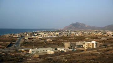 تظهر الصورة منظرًا خلابًا لبلدة ساحلية تطل على البحر. يُظهر المشهد منازل مبنية بشكل متفرق، بعضها حديث والآخر تقليدي، مع شوارع تمتد بين المباني. في الخلفية، يمكن رؤية سلسلة جبال، بينما يمتد البحر الفسيح على الجهة اليسرى. السماء صافية وتبدو أشعة الشمس مشرقة، مما يضفي شعورًا بالهدوء والجمال الطبيعي على المكان.