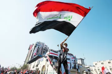 L'image montre une personne agitant un drapeau irakien. Le drapeau, aux couleurs rouge, blanc, noir et vert, est bien visible. La scène se déroule probablement lors d'une manifestation ou d'un événement public, avec des bâtiments en arrière-plan décorés de drapeaux. L'atmosphère semble dynamique, reflétant un sentiment de patriotisme et de rassemblement.