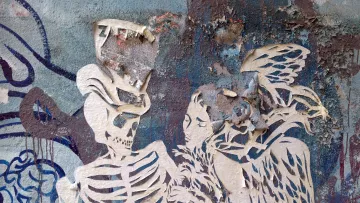 The image features a graffiti-style artwork on a textured wall. It depicts a skeletal figure, a skeleton, in an intimate pose with a woman who seems to have angelic features, including wings. The artwork appears to be layered, with parts of the paper or paint peeling away, adding a weathered and distressed look. The background consists of blue and gray tones, enhancing the overall atmosphere of the piece. The combination of the skeleton and the woman creates a striking contrast, possibly symbolizing themes of life and death, or love and loss.