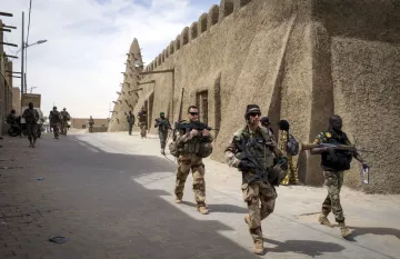 تظهر الصورة جنوداً يسيرون في شوارع منطقة تاريخية. المباني في الخلفية تبدو مصنوعة من الطين، وتمتاز بتصميم تقليدي. الجنود يحملون أسلحة ويبدو عليهم التركيز والحذر. يتواجد أيضاً شخصيات أخرى في الخلفية، مما يدل على وجود نشاطات متعددة في المنطقة. السماء تبدو صافية مع بعض السحب.