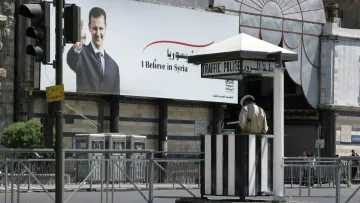 L'immagine mostra una cabina di polizia del traffico situata in una strada. In secondo piano, c'è un grande manifesto che ritrae un uomo che saluta, con il testo in arabo e italiano che dice "I Believe in Syria". La scena è animata ma appare tranquilla, con poche auto visibili e un'illuminazione chiara. Il contrasto tra la cabina di polizia e il manifesto può suggerire elementi di sicurezza e comunicazione politica.