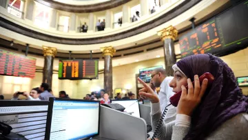 تظهر الصورة مشهداً من داخل سوق الأسهم، حيث يوجد عدد من الأشخاص يتعاملون مع شاشات تعرض الأسعار والبيانات المالية. في المقدمة، توجد امرأة ترتدي حجاباً، وهي تتحدث عبر الهاتف. يبدو أنها مشغولة بالعمل، حيث تشير بإحدى يديها. الخلفية تحتوي على عدة أشخاص آخرين وشاشات تعرض معلومات السوق. الأجواء توحي بالجدية والنشاط المالي.