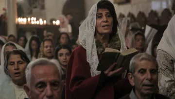 L'immagine mostra un gruppo di persone durante una cerimonia, probabilmente in una chiesa. Una donna al centro, che indossa un velo bianco e un abito rosso, tiene in mano un libro e sembra partecipare attivamente alla celebrazione. Intorno a lei ci sono altre persone, alcune delle quali appaiono in preghiera o cantando. L'atmosfera è solenne, con un gioco di luci morbide e candele accese sullo sfondo che creano un'illuminazione calda e accogliente.