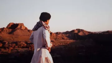 در این تصویر، فردی با لباس سنتی و شال بر دوش، به سمت یک چشم‌انداز طبیعی نگاه می‌کند. زمینه تصویر شامل کوه‌های بلند و دشت‌های وسیع است که نور ملایم غروب خورشید بر آنها تابیده شده است. رنگ‌های گرم غروب و سایه‌های بلند کوه‌ها زیبایی خاصی به صحنه داده‌اند. احساس آرامش و سکوت در فضای تصویر به وضوح حس می‌شود.